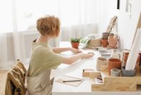 aap-how-to-create-a-craft-room-on-a-small-budget-1791727224 A woman sits in a cozy workspace crafting with flower pots and supplies.