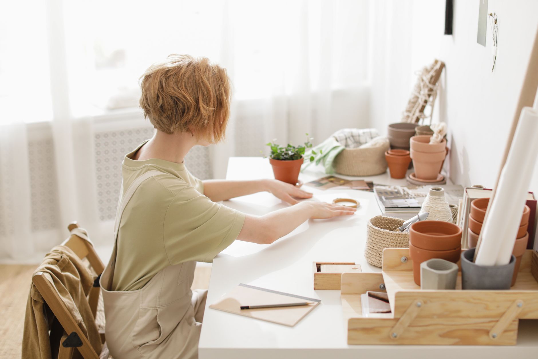 A woman sits in a cozy workspace crafting with flower pots and supplies.