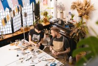 Two artisans creating jewelry pieces in a sunlit studio filled with plants and tools.