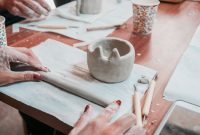 Close-up of hands sculpting pottery with tools in an indoor workshop setting.