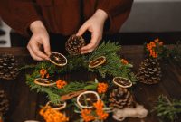 aap-seasonal-wreath-making-ideas-for-fall-1791813622 Hands arranging pine cones and dried oranges on a Christmas wreath.