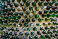 aap-upcycled-wine-bottle-craft-projects-1791640820 Creative display of empty glass bottles embedded in a wall, forming a colorful pattern.